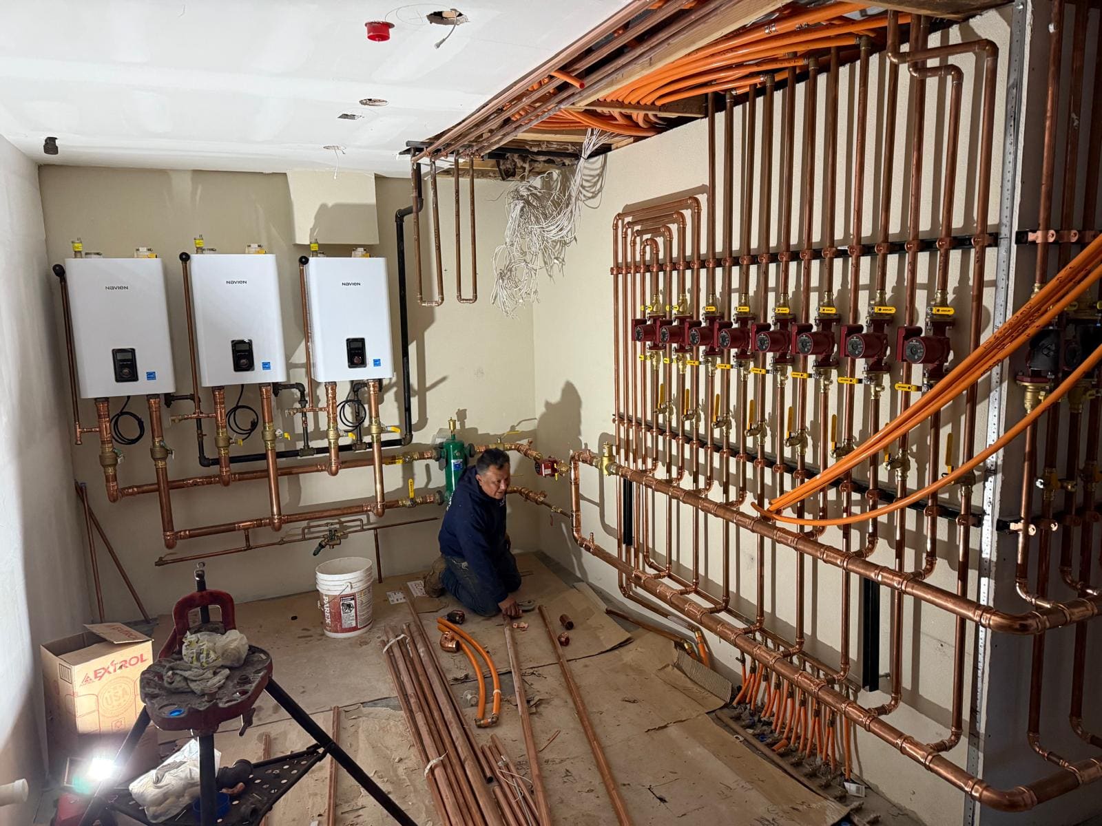 PLUMBON24 plumber at work in a mechanical room with three wall-hung boilers and a copper heating manifold